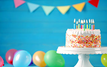Delicious birthday cake on a white tray with burning candles and colorful balloons on table